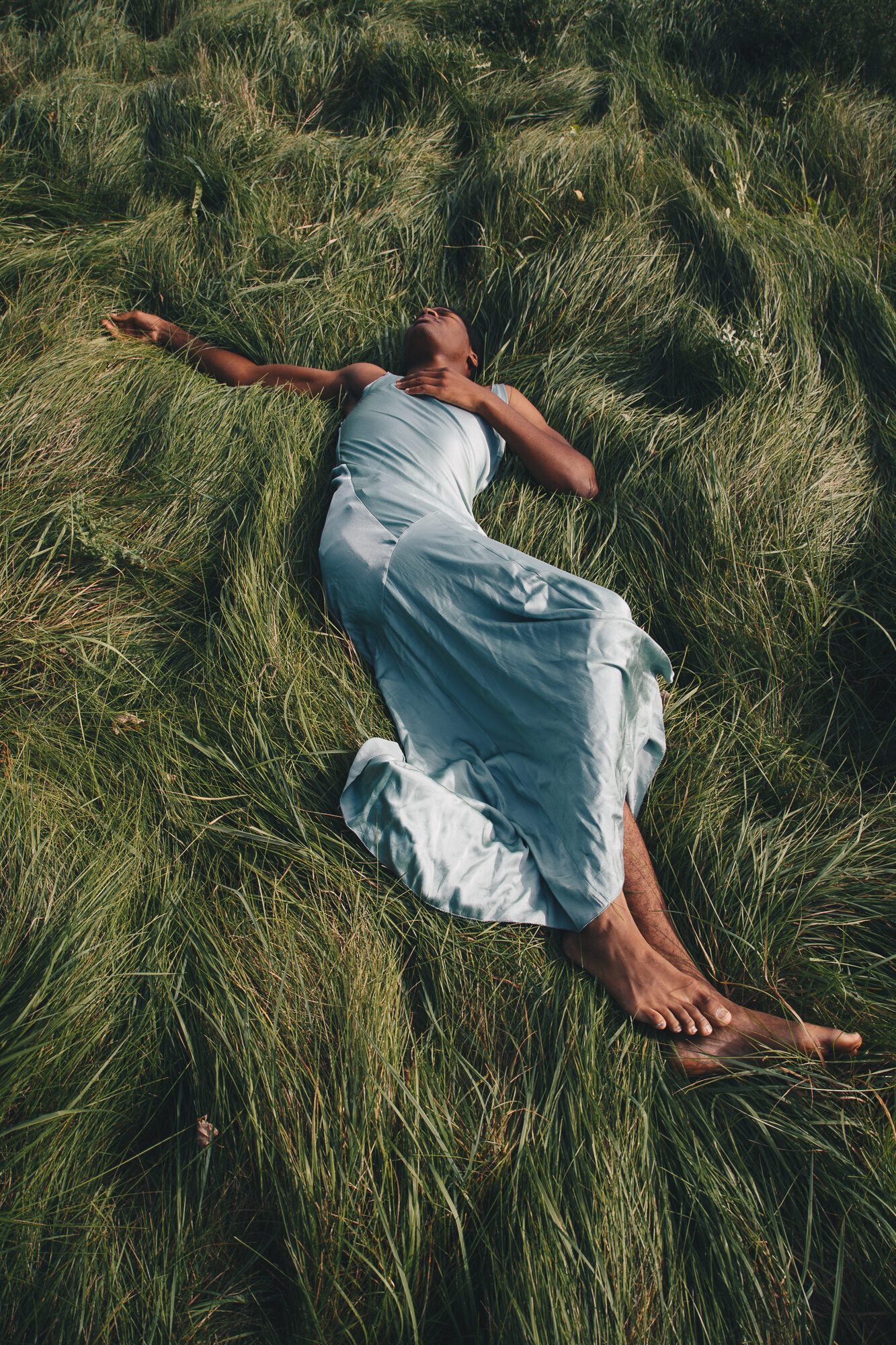 Person in a pale blue dress lying in tall grass, editorial portrait photography outdoors