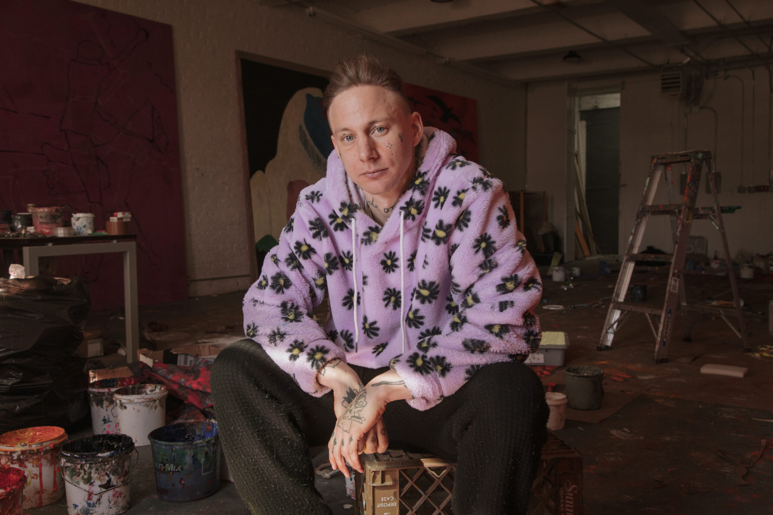 Man seated indoors wearing a patterned shirt, warm ambient light and candid expression.