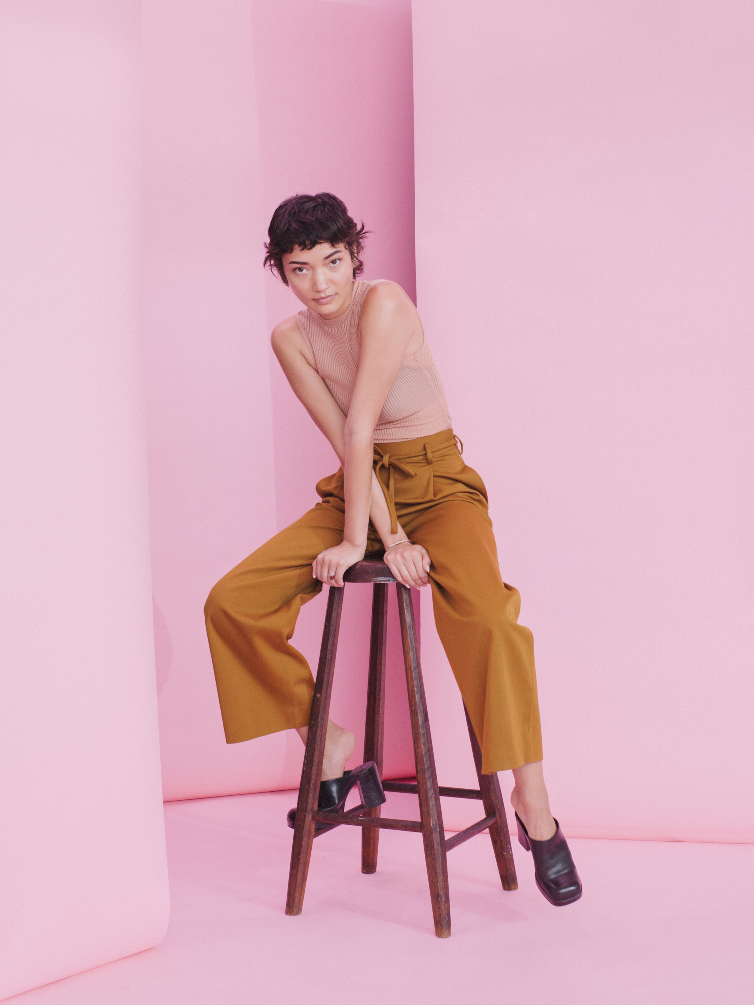 Fashion portrait of a person posed on a stool in a pink studio set wearing tan pants.