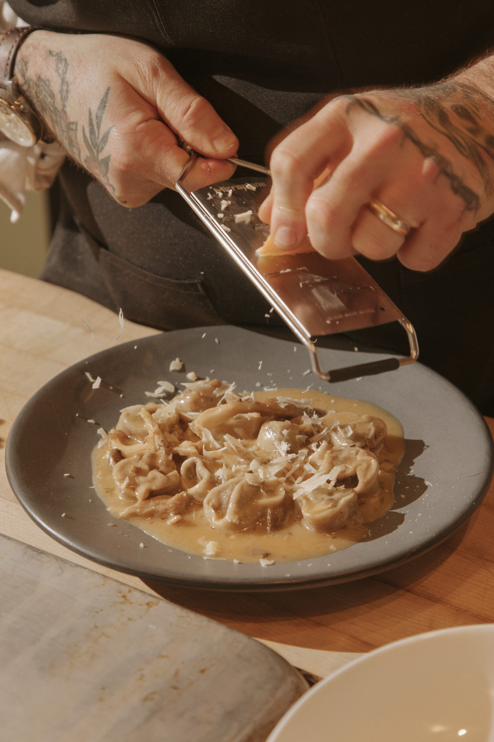Close-up food photo of pasta being finished with grated cheese, warm kitchen lighting.