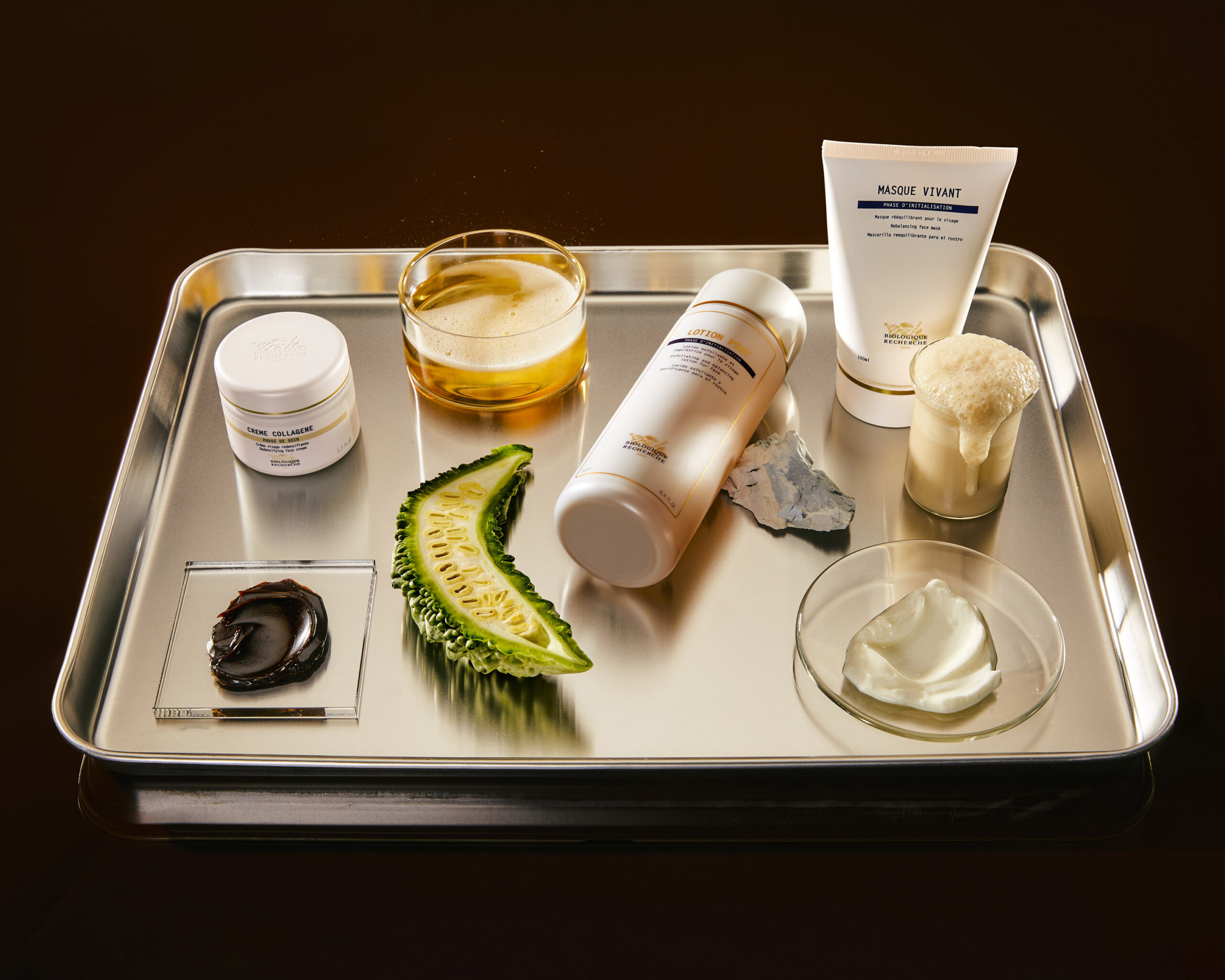 Skincare and wellness products arranged on a metal tray with avocado and glassware