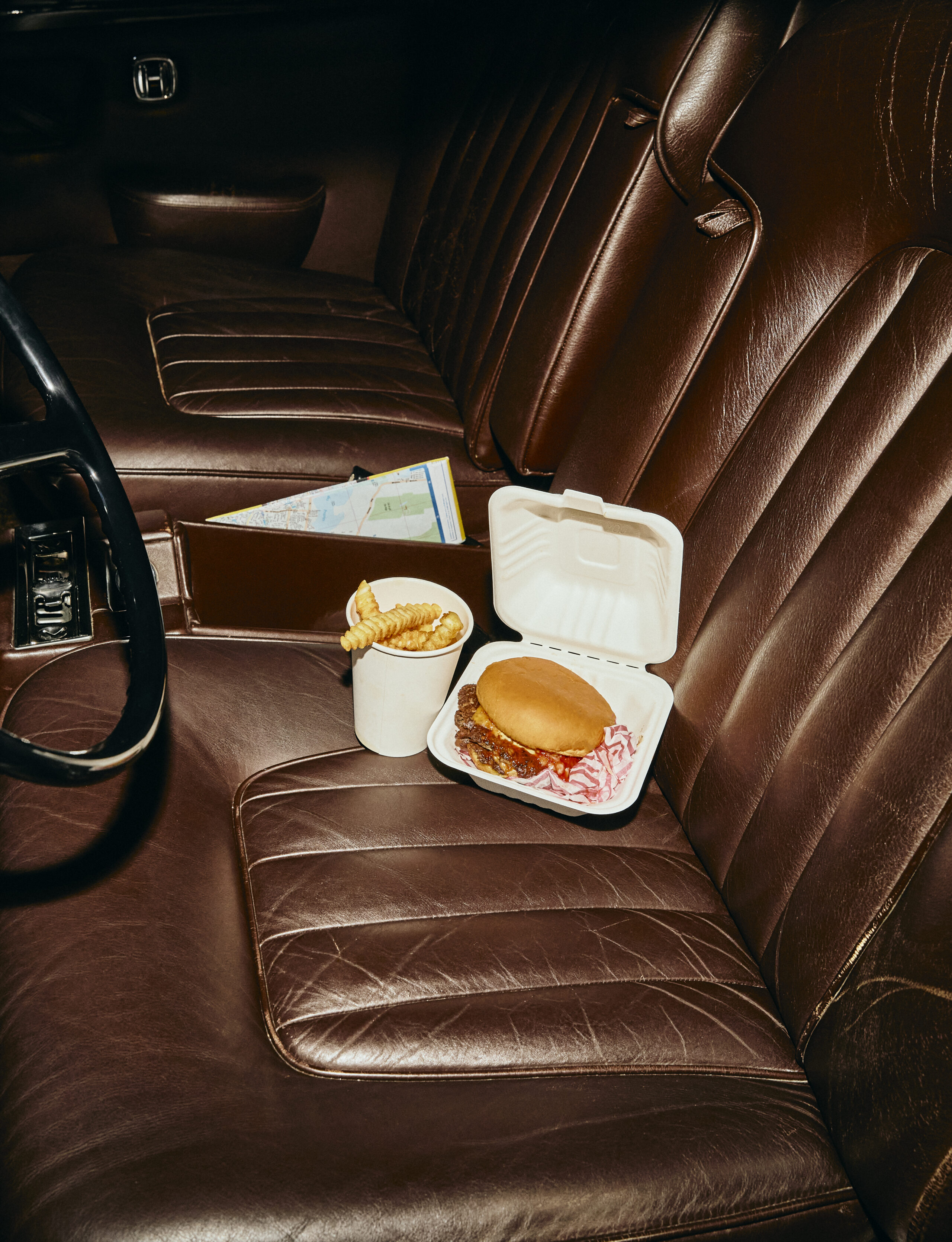 Takeout burger, fries, and drink arranged on a brown leather car seat in a retro interior