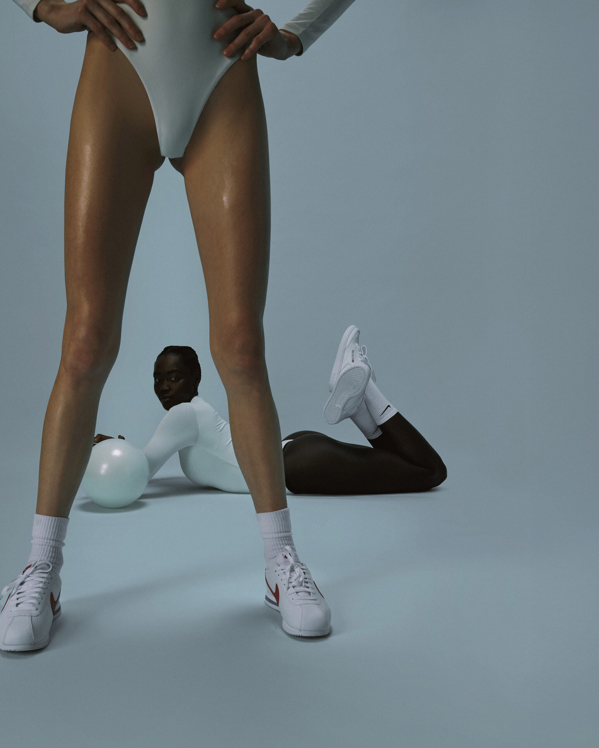 Fitness image with standing athlete’s legs in foreground and another person posed on the floor in a studio