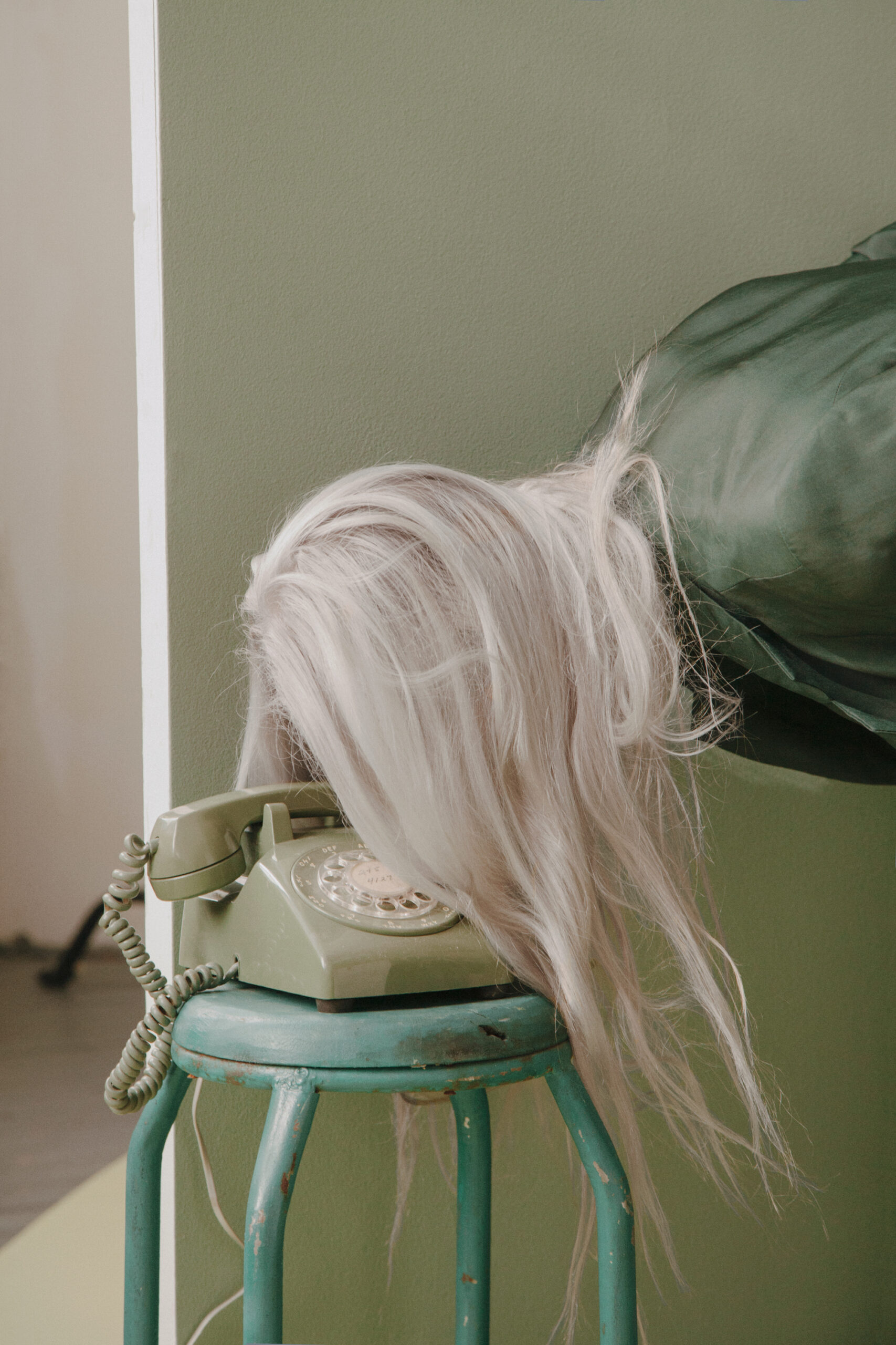 Blonde person leaning forward near a green vintage phone on a stool